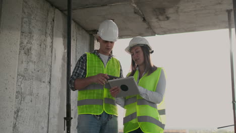 Engineers-designers-a-man-and-a-woman-standing-on-the-roof-of-a-building-under-construction-and-discuss-the-plan-and-progress-of-construction-using-a-tablet-and-mobile-phone.-Modern-builders-discuss-the-infrastructure-of-the-building-and-the-surrounding-area